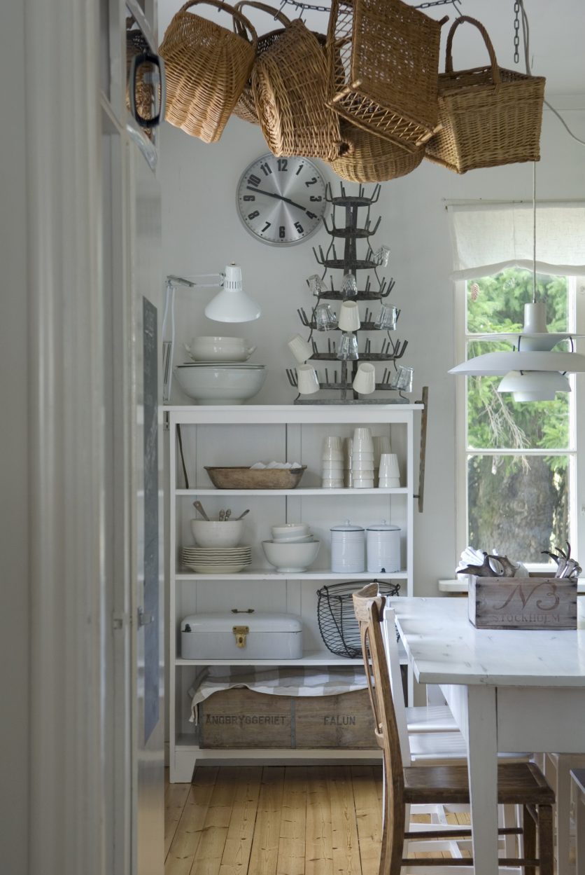 Corner of dining room with white dining set, baskets hanging from ceiling and white-painted, open-fronted shelves of crockery ag